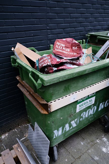 A green wheelie bin filled with several red and black sacks of garden or construction waste, positioned on a paved surface next to a dark brick wall. The sacks are marked with labels indicating they c
